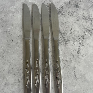 May include: Four stainless steel butter knives with leaf pattern handles. The knives are arranged vertically against a mottled grey background. The blades are smooth and the handles have a decorative leaf design.