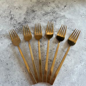 May include: Six gold-coloured forks arranged in a fan shape. Each fork has four tines and a long, slender handle. The forks are made of a shiny, metallic material, set against a mottled grey background.
