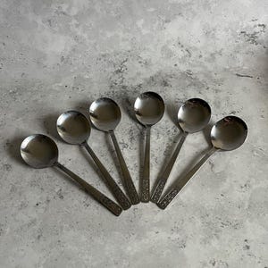 May include: Six stainless steel soup spoons arranged in a fan shape. Each spoon has a round bowl and a long handle with a decorative pattern near the base. The spoons are set against a mottled grey background.
