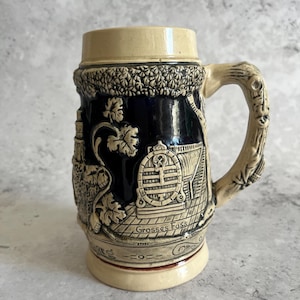 May include: A vintage ceramic beer stein with a cream-coloured top and base, and a dark blue body. The body features embossed scenes of a barrel, buildings, and foliage. The handle is designed to resemble a tree branch, and the text "Grosses Fass" is visible.