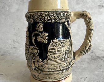 May include: A vintage ceramic beer stein with a cream-coloured top and base, and a dark blue body. The body features embossed scenes of a barrel, buildings, and foliage. The handle is designed to resemble a tree branch, and the text "Grosses Fass" is visible.