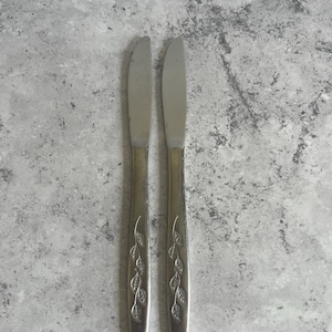 May include: Two stainless steel butter knives with leaf pattern handles. The knives are arranged vertically against a mottled grey background. The blades are smooth and the handles feature a decorative leaf design.