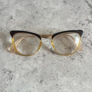May include: Vintage cat-eye eyeglasses with black upper rims and gold-toned metal frames. The lenses are clear, and the lower rims are a light yellow color. The glasses have a classic, retro style.