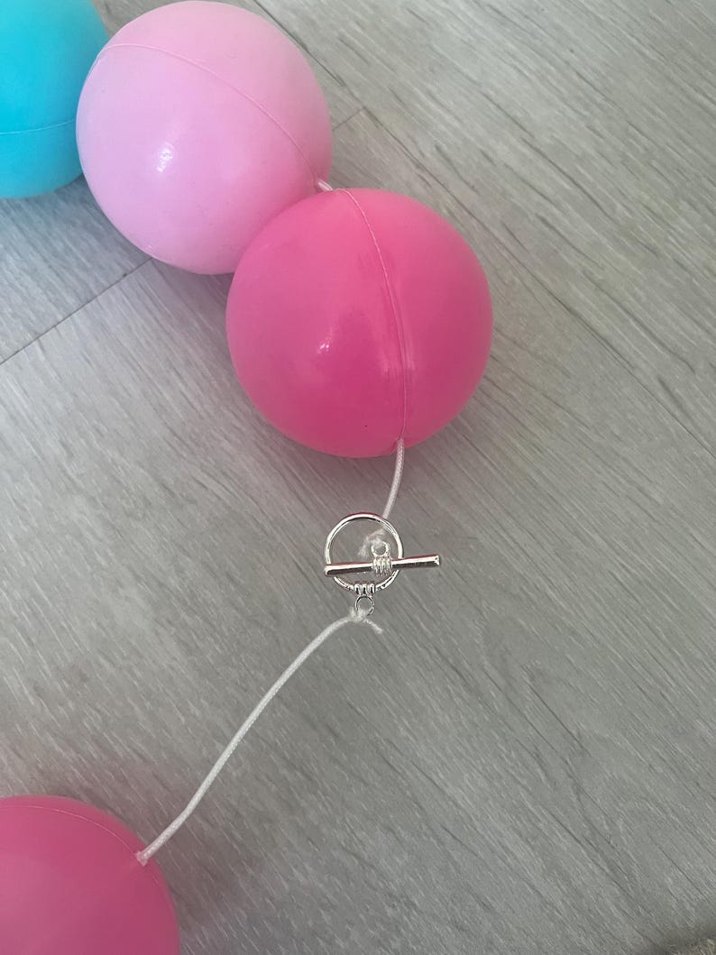 K&ouml;nnte beinhalten: Eine Girlande aus rosa und blauen Luftballons mit einem silbernen Toggle-Verschluss. Die Ballons sind mit wei&szlig;em Garn verbunden.