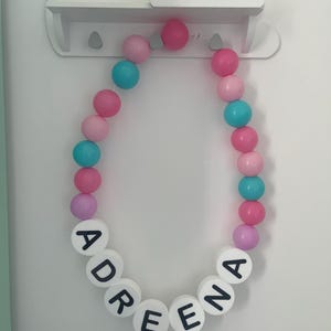 K&ouml;nnte beinhalten: Ein wei&szlig;es Wolkenregal mit einer rosa-blauen Perlenkette, die den Namen "Adreena" buchstabiert.