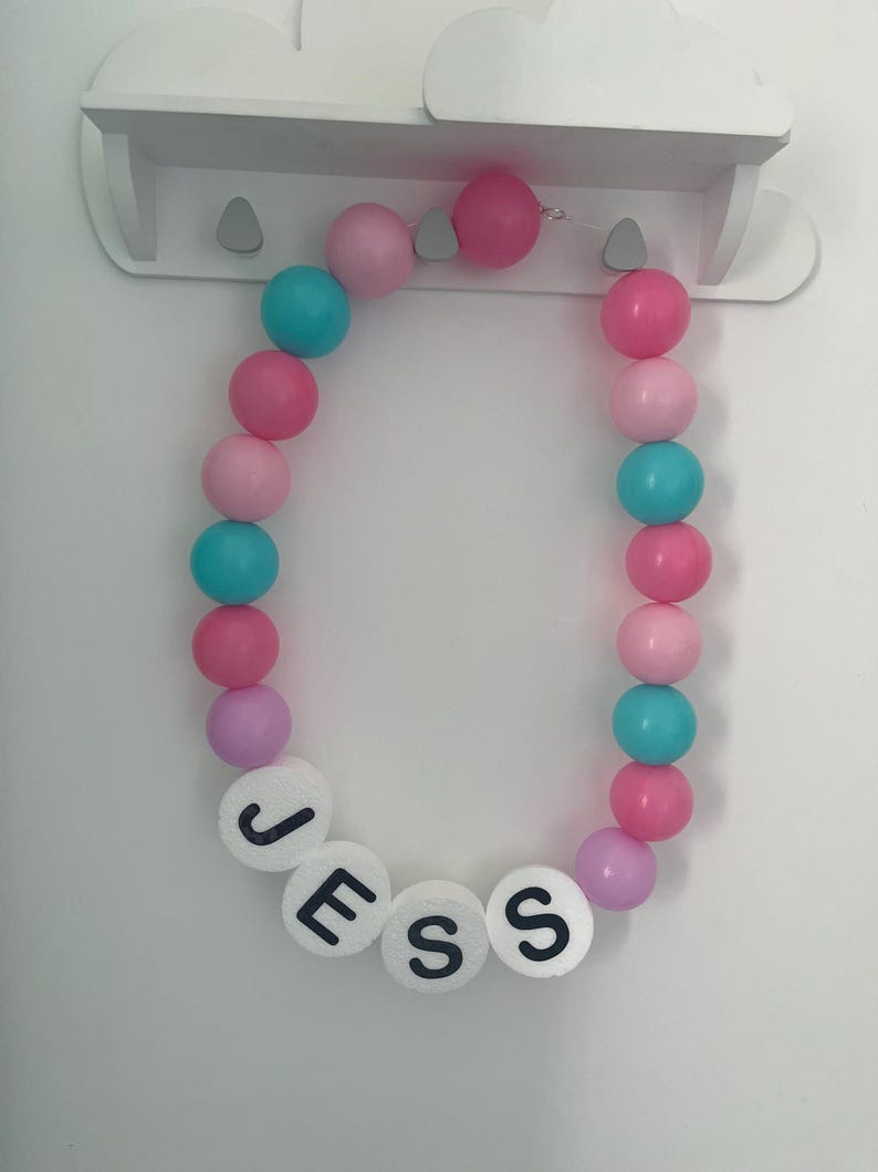 K&ouml;nnte beinhalten: Eine rosa und blaue Perlenkette mit dem Namen "JESS" in wei&szlig;en Buchstaben.