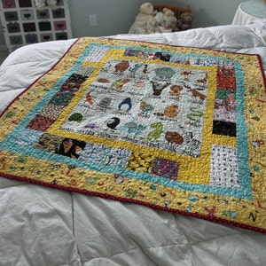 May include: A yellow and red quilted baby blanket with a colorful alphabet design featuring animals and letters. The blanket has a blue and white border with a yellow and red polka dot trim.