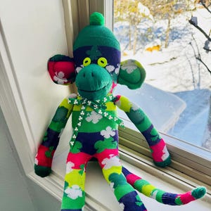 May include: A green, blue, pink, yellow, and white sock monkey with a green hat and shamrock patterns. The monkey is sitting on a windowsill.