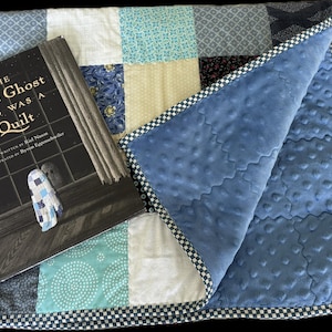May include: A patchwork quilt featuring various blue, white, and cream fabric squares with a textured blue minky backing and a checkered border, displayed alongside a book titled The Little Ghost Who Was a Quilt by Riel Nason.