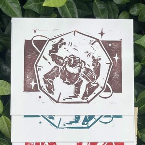 Puede incluir: Una pila de cinco impresiones rectangulares de papel con una ilustración estilo linograbado de una criatura dentro de una forma geométrica. Las impresiones se muestran en varios colores de tinta sobre papel blanco, tostado y crema.