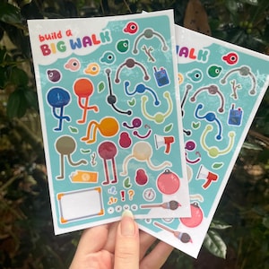 May include: Two rectangular sticker sheets with a light blue background featuring the text build a BIG WALK. The sheets contain various colorful cartoon character parts, including round heads, thin limbs, small accessories like a megaphone, a compass, and a paintbrush, along with a blank white rectangular label.