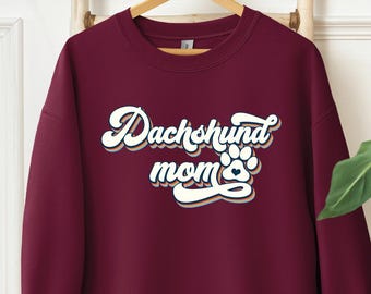 May include: Maroon sweatshirt with the text "Dachshund mom" in a retro font, featuring a paw print and heart design. The long-sleeved crewneck top is hanging on a wooden ladder, showcasing a casual style.