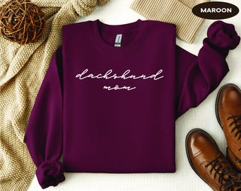May include: A maroon sweatshirt with the words "dachshund mom" in white script. The long-sleeved crew neck shirt is displayed with brown leather boots and a beige knit hat. A decorative woven ball and a beige knit blanket are also in the image.