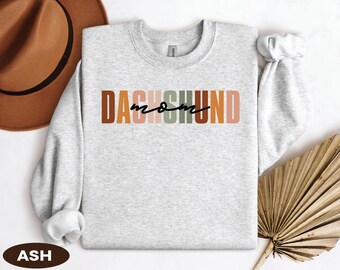 May include: A light gray sweatshirt with the text "DACHSHUND mom" in a colorful, layered design. The word "DACHSHUND" is in brown, orange, and green blocks, with "mom" in a handwritten font. A brown hat and dried plant are in the background.