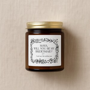 May include: A brown glass jar with a gold lid. The jar features a white label with the text "MAYA, WILL YOU BE MY BRIDESMAID?" in black, framed by floral illustrations. The text "I can't say I do without you" is below.