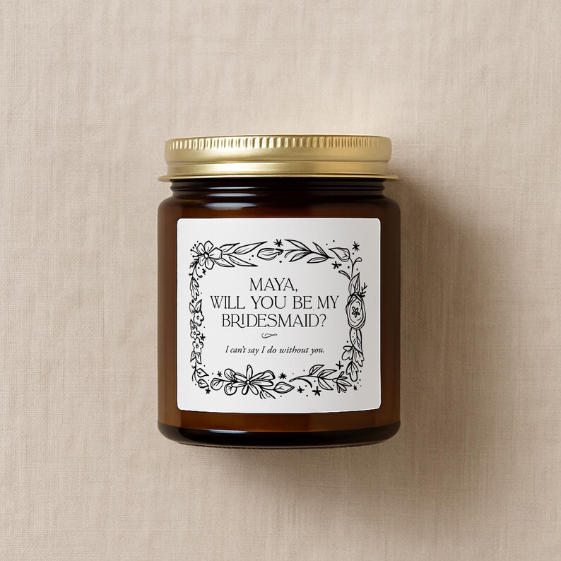 May include: A brown glass jar with a gold lid. The jar features a white label with the text "MAYA, WILL YOU BE MY BRIDESMAID?" in black, framed by floral illustrations. The text "I can't say I do without you" is below.