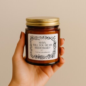 May include: A brown glass candle jar with a gold lid. The label reads "MAYA, WILL YOU BE MY BRIDESMAID? I can't say I do without you." The label is surrounded by a floral design. The jar is held in a hand.
