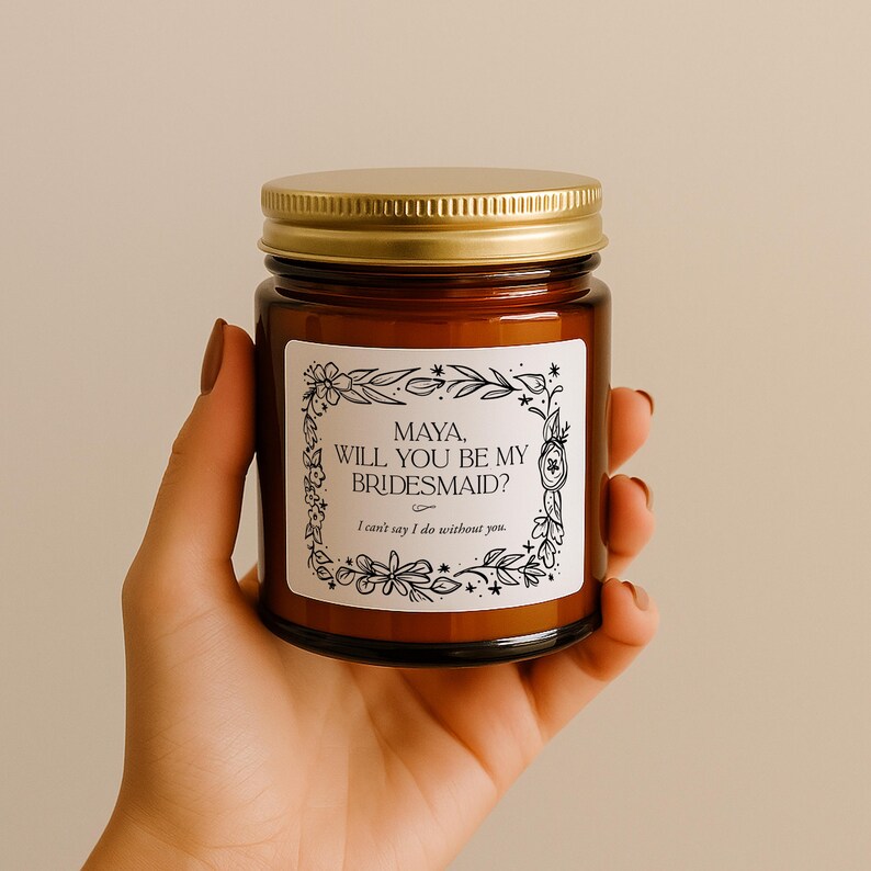 May include: A brown glass candle jar with a gold lid. The label reads "MAYA, WILL YOU BE MY BRIDESMAID? I can't say I do without you." The label is surrounded by a floral design. The jar is held in a hand.