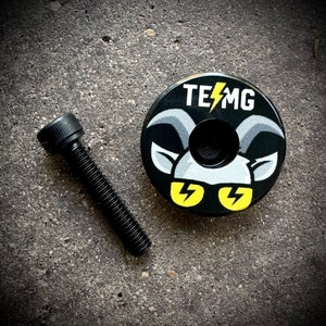 Puede incluir: Tapa de potencia de bicicleta negra con un diseño de cabra blanca y rayos amarillos. El texto "TE⚡️MG" está impreso en la tapa.