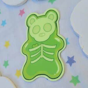 May include: A green gummy bear sticker with a skeleton inside. The bear is outlined in black and has a heart on its chest.