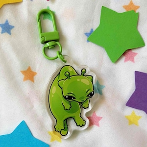 Puede incluir: Llavero de gato extraterrestre verde con un cierre de metal verde. El llavero está hecho de acrílico transparente y presenta un gato extraterrestre de dibujos animados con ojos grandes y una boca pequeña. El gato extraterrestre está sentado sobre sus patas traseras con las patas delanteras levantadas.