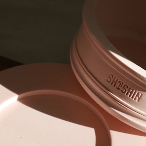 Op de afbeelding: Een roze keramische plantenpot met het woord "SHOSHIN" in reliëf aan de zijkant. De pot heeft een bijpassende schotel.