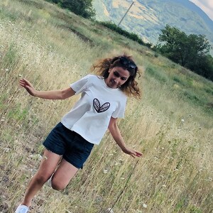 May include: White t-shirt with a black leaf design, paired with dark blue denim shorts and white sneakers. The person is in a grassy field with mountains in the background. Sunglasses are on top of the head.