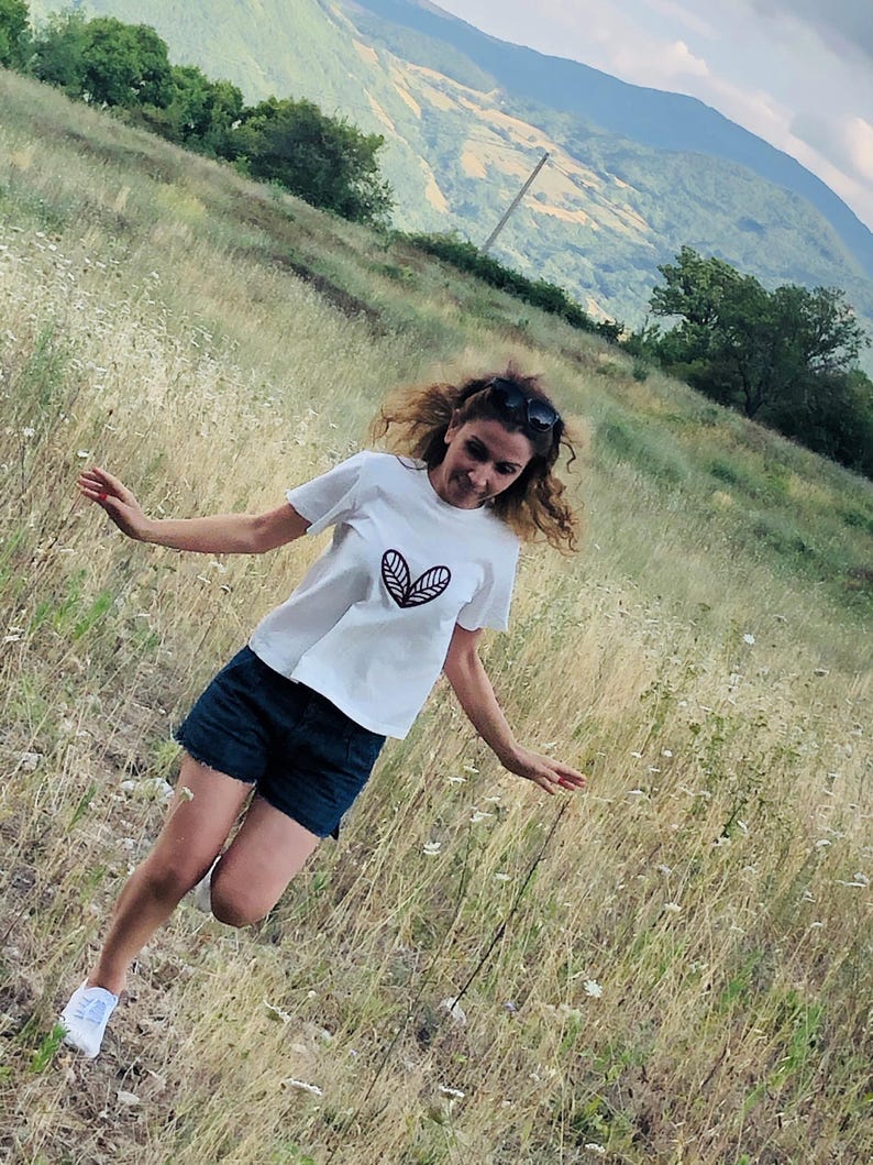 May include: White t-shirt with a black leaf design, paired with dark blue denim shorts and white sneakers. The person is in a grassy field with mountains in the background. Sunglasses are on top of the head.