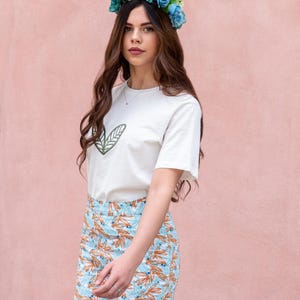 May include: A woman wearing a white t-shirt with a green leaf design, paired with patterned pants in shades of blue and orange. She is also wearing a blue flower crown. The background is a solid pink color.