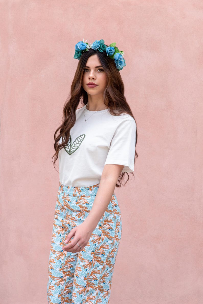 May include: A woman wearing a white t-shirt with a green leaf design, paired with patterned pants in shades of blue and orange. She is also wearing a blue flower crown. The background is a solid pink color.