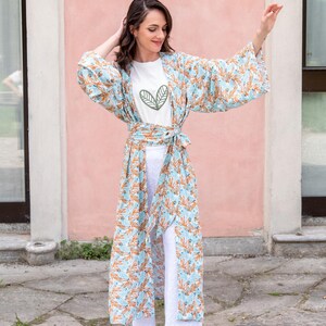 May include: A woman wearing a light blue kimono with an orange floral pattern over a white t-shirt and white pants. The t-shirt has a green leaf design. The kimono is tied at the waist with a matching belt.