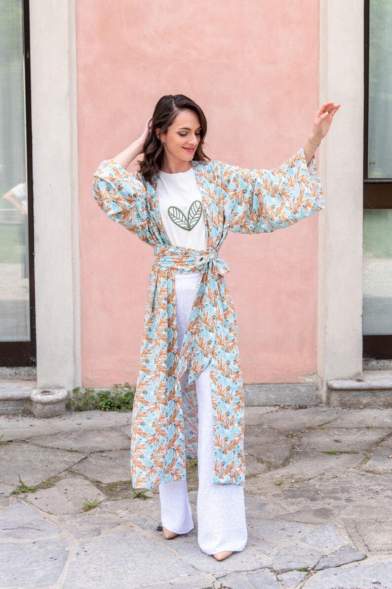 May include: A woman wearing a light blue kimono with an orange floral pattern over a white t-shirt and white pants. The t-shirt has a green leaf design. The kimono is tied at the waist with a matching belt.