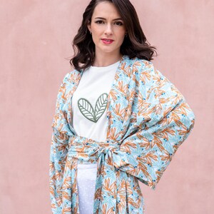 May include: A woman wearing a white t-shirt with a green leaf design and a light blue and orange floral print kimono. The kimono is tied at the waist. The background is a solid pink color.