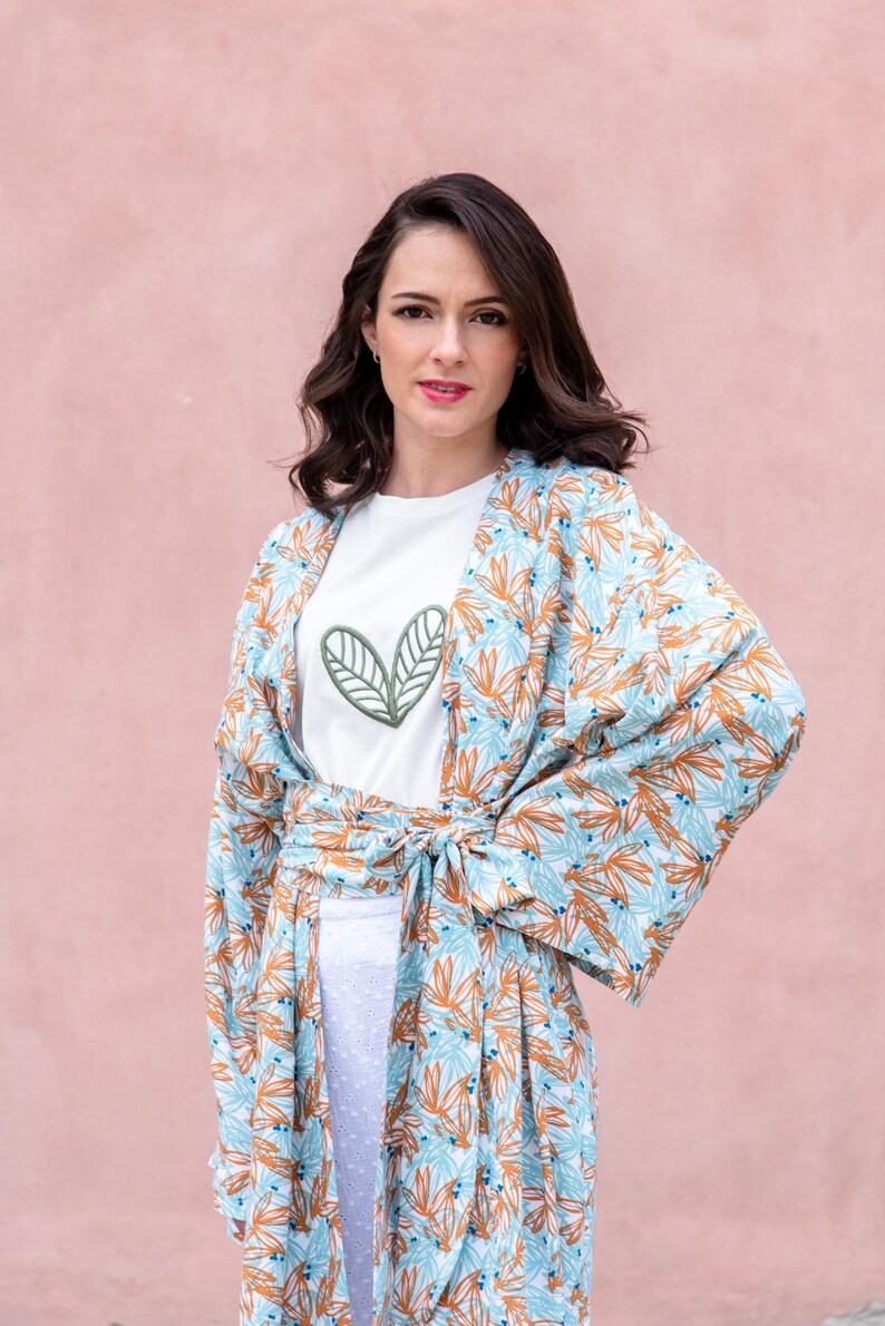 May include: A woman wearing a white t-shirt with a green leaf design and a light blue and orange floral print kimono. The kimono is tied at the waist. The background is a solid pink color.