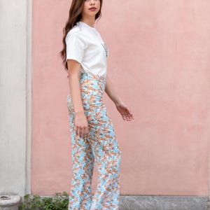 May include: A woman wearing a white t-shirt and light blue floral print pants. The pants have an orange leaf pattern. She is also wearing a blue flower crown and black sandals. The background is a pink wall.