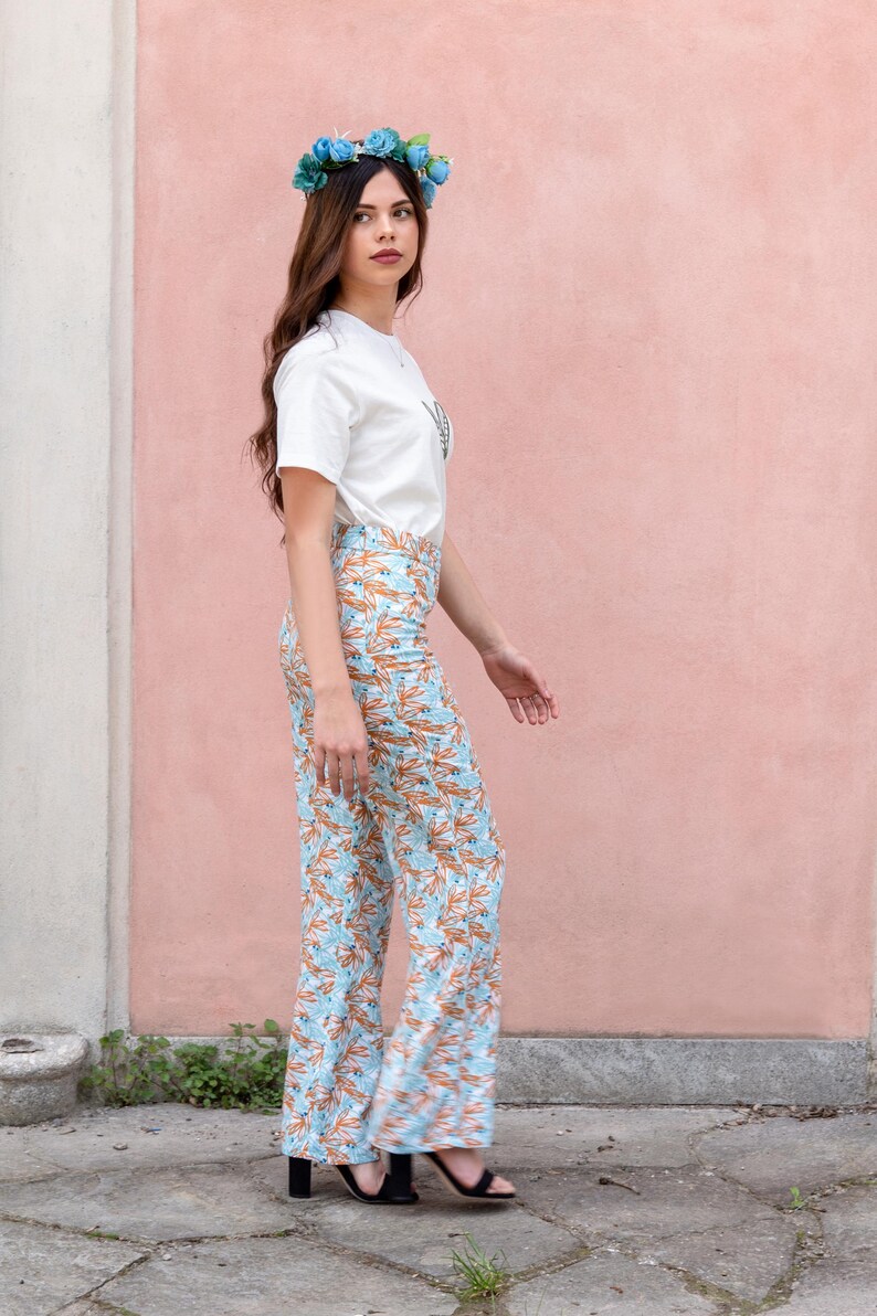 May include: A woman wearing a white t-shirt and light blue floral print pants. The pants have an orange leaf pattern. She is also wearing a blue flower crown and black sandals. The background is a pink wall.