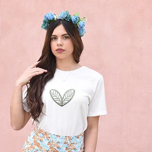 May include: White t-shirt with a green leaf design. The model is wearing a blue flower crown. The background is a solid pink color.