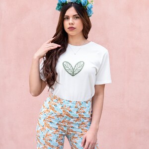 May include: A white t-shirt with a green leaf design, paired with patterned pants in shades of blue, orange, and white. A blue floral crown adorns the head. The background is a solid pink.