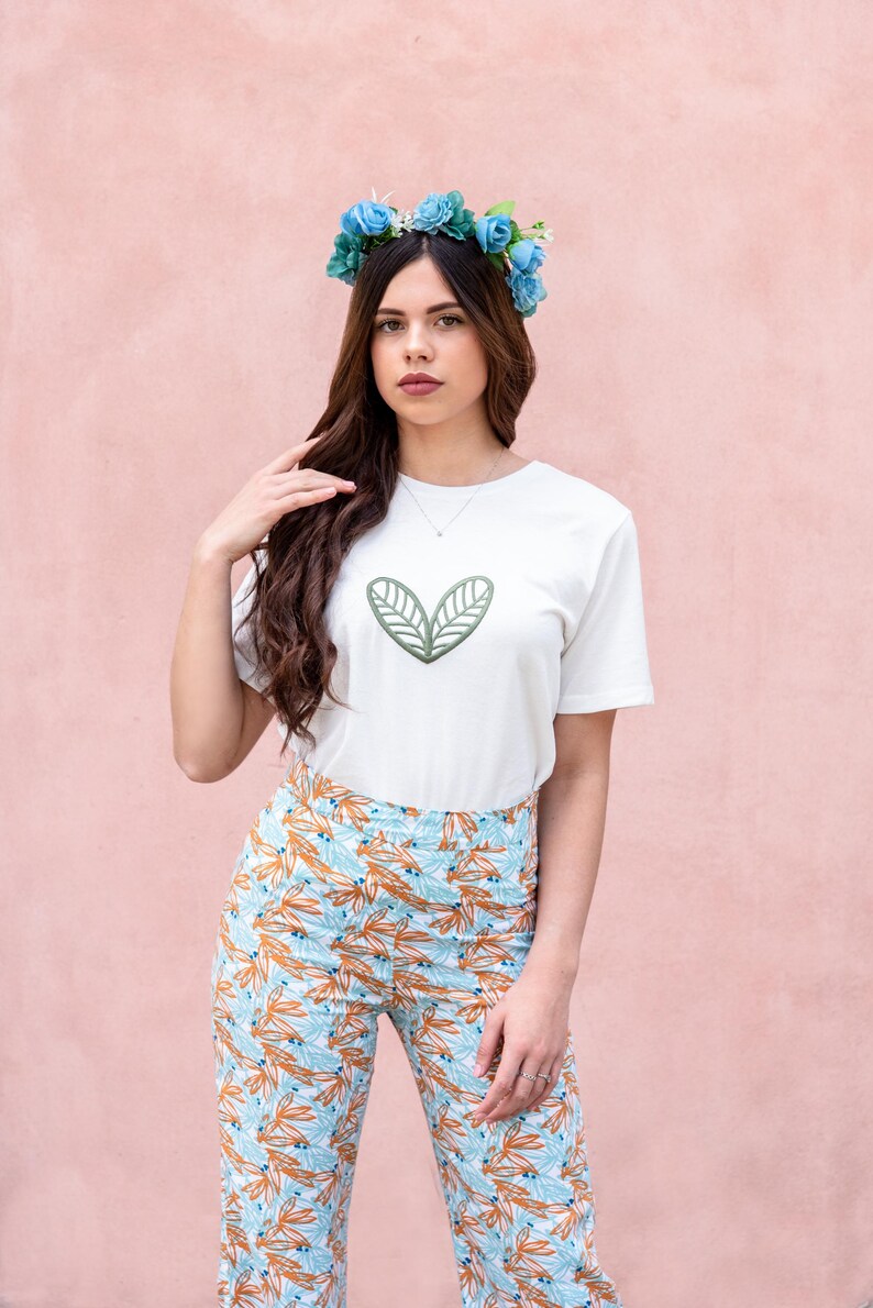 May include: A white t-shirt with a green leaf design, paired with patterned pants in shades of blue, orange, and white. A blue floral crown adorns the head. The background is a solid pink.