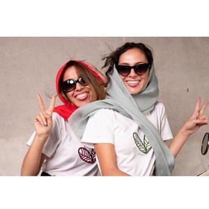 May include: Two people wearing sunglasses and white t-shirts with leaf designs. One person is wearing a red headscarf, the other a light green headscarf. Both are smiling and making peace signs.