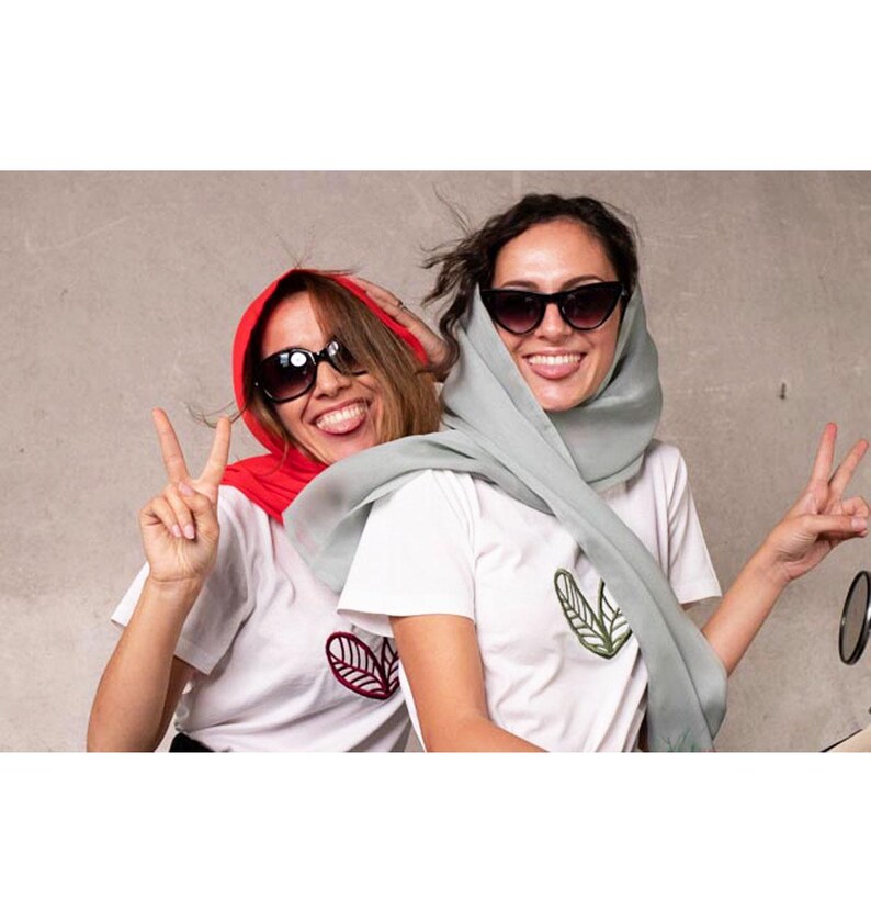 May include: Two people wearing sunglasses and white t-shirts with leaf designs. One person is wearing a red headscarf, the other a light green headscarf. Both are smiling and making peace signs.