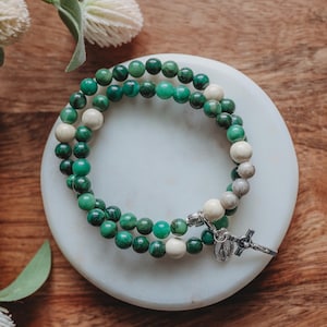 May include: A beaded bracelet featuring green and off-white round beads. The bracelet is adorned with a silver-colored cross pendant and a small charm. The beads are arranged in a double strand, resting on a white, circular surface.