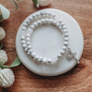 Puede incluir: Una pulsera de cuentas blancas y grises con un dije de cruz plateada descansa sobre una superficie de mármol blanco. La pulsera está hecha de cuentas redondas veteadas y se exhibe sobre un fondo de madera con flores blancas.