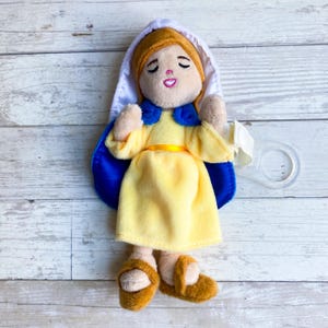 May include: A plush doll depicting a religious figure, featuring a yellow dress, blue cape, and white head covering. The doll has closed eyes and a smiling expression. It includes a clear plastic loop.
