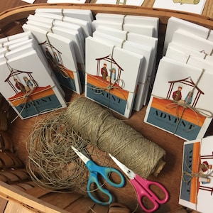 May include: A basket filled with stacks of white cards tied with twine. The cards feature a nativity scene illustration and the text "Legacy Church Advent Family Moments Cards". A roll of twine and a pair of scissors are in the bottom of the basket.
