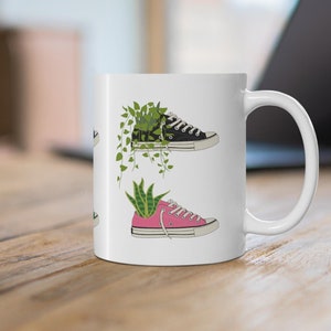 Puede incluir: Taza de cerámica blanca con un diseño de zapatillas negras y rosas. Cada zapatilla tiene una planta verde que crece fuera de ella.
