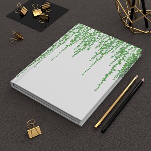May include: A white notebook with a green leafy pattern on the cover. The notebook is closed and lying flat on a dark gray surface. Two pencils, one gold and one black, are lying next to the notebook.