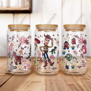 May include: Three clear glass tumblers with bamboo lids and straws. Each tumbler features a different Toy Story character design, including Woody, Buzz Lightyear, and others, surrounded by floral patterns. The tumblers are set on a wooden surface.