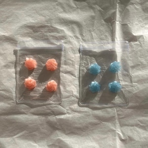 Op de afbeelding: Twee doorzichtige plastic zakjes met rits, elk met vier kleine, kleurrijke, roosvormige snoepjes. Eén zakje bevat roze snoepjes, het andere blauwe snoepjes.