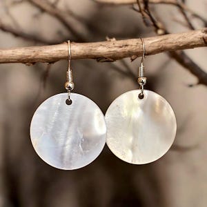 May include: A pair of round, iridescent white shell earrings. Each earring features a circular shell pendant suspended from a silver-colored hook. The earrings are hanging from a brown branch, with a blurred background.